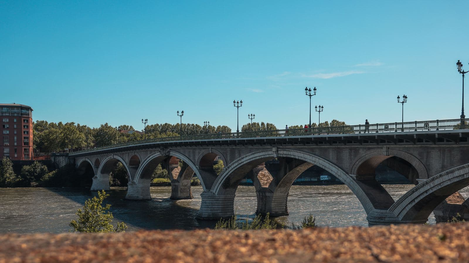 A stone and steel arch bridge crossing a river, with a city visible on the far bank